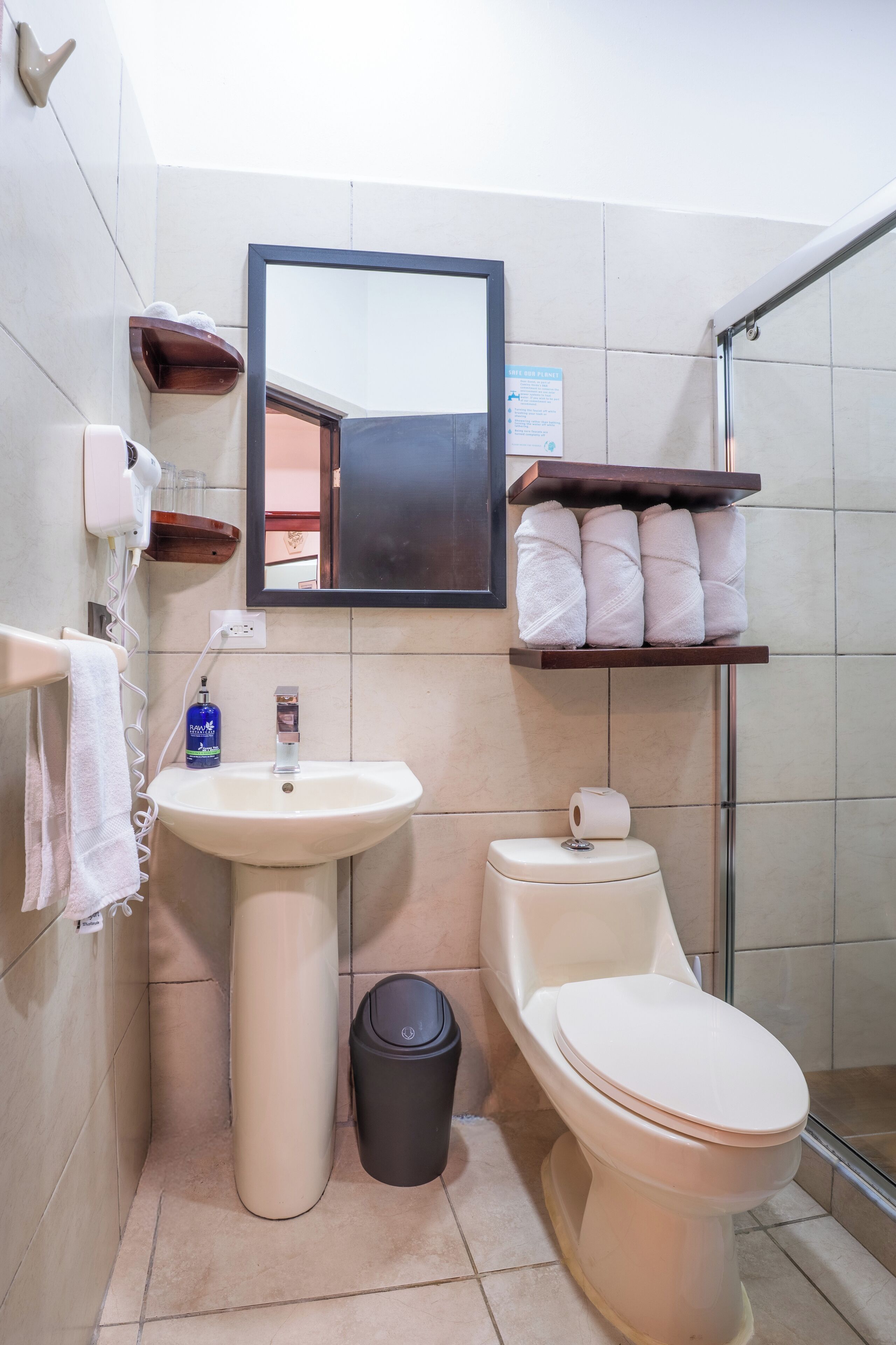 A bathroom with a sink, toilet, and a mirror. There are towels on a shelf and a trash bin.
