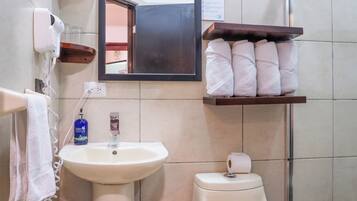 Baño con lavabo, inodoro y espejo. Hay toallas en un estante y un cesto de basura.