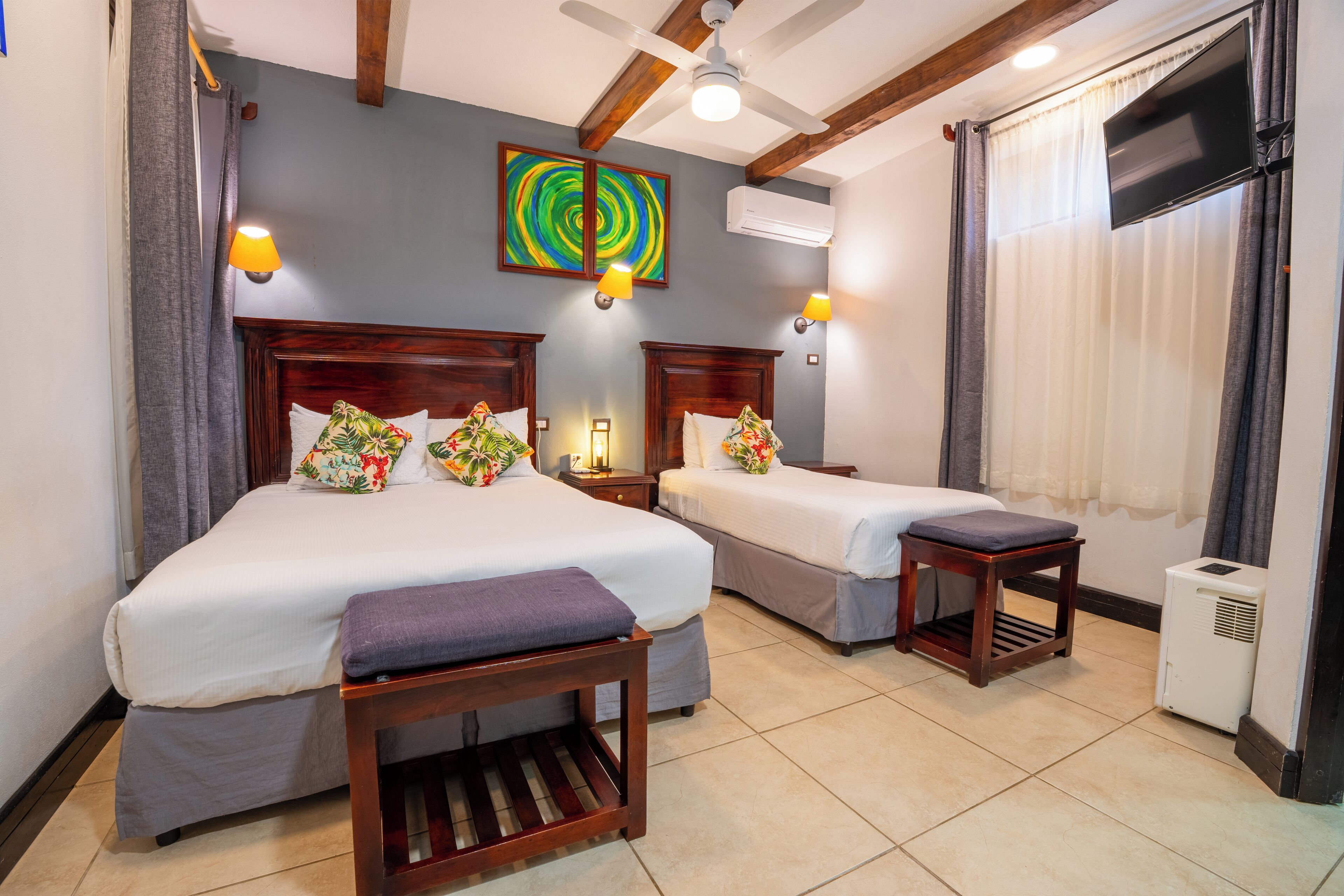 A hotel room with two beds, a ceiling fan, a TV mounted on the wall, and a window with curtains.