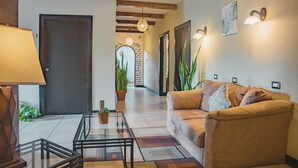 A living room with a sofa, a glass coffee table, and a lamp. There is a door and a hallway leading to another room.