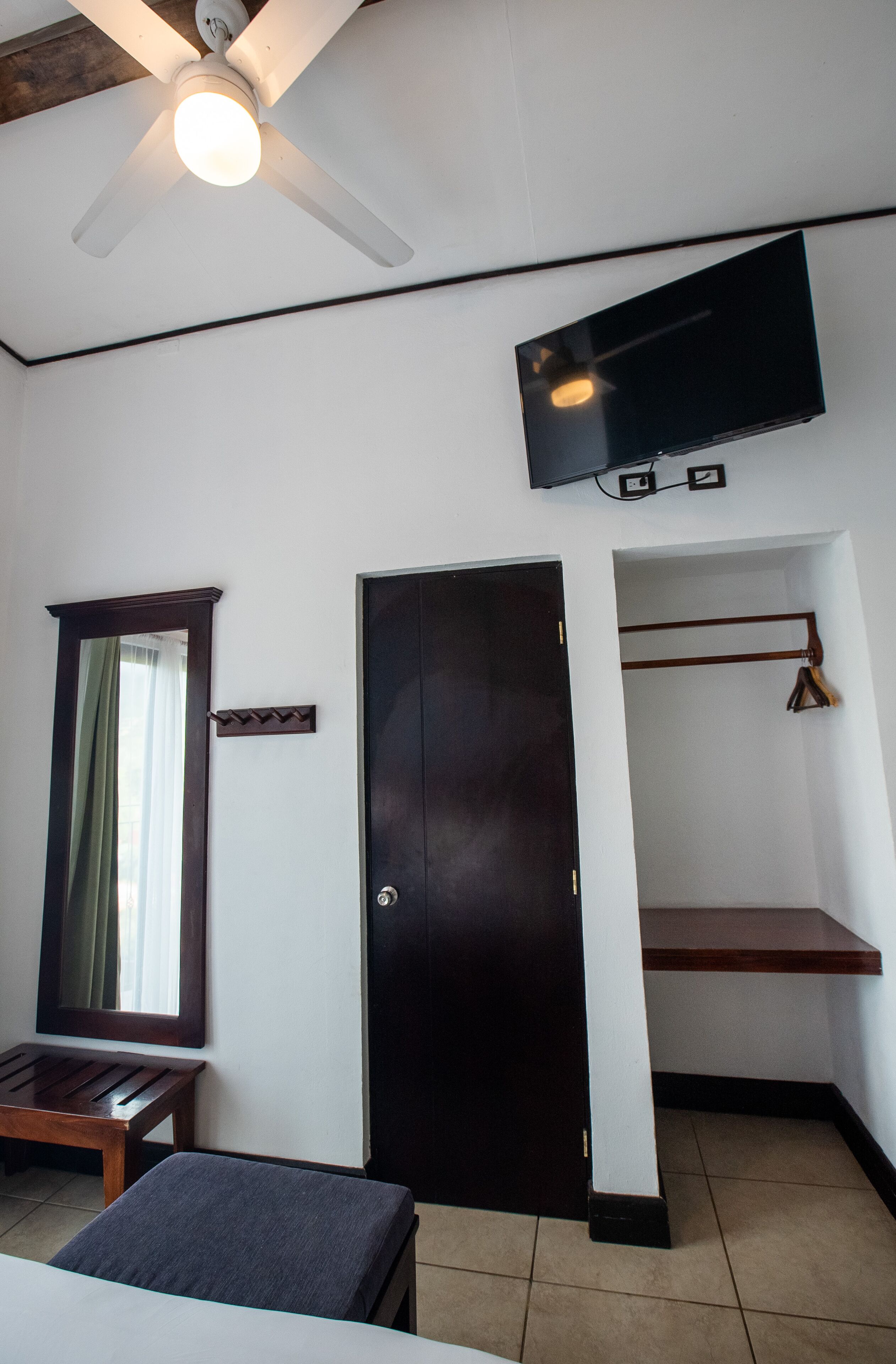 A room with a ceiling fan, a flat-screen TV mounted on the wall, a wooden bench, a door, and a small wooden shelf.