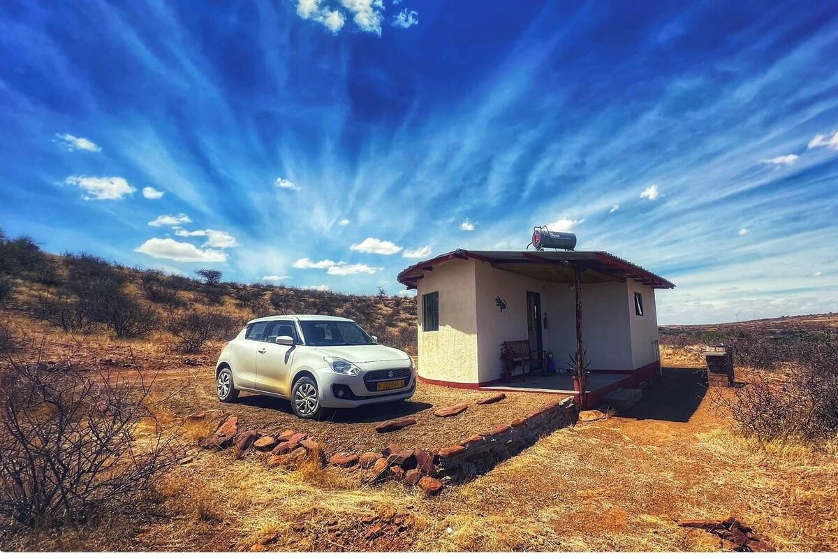 A small house with a car parked in front, surrounded by dry grass and shrubs.
