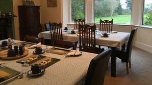 Une salle à manger avec une table dressée pour quatre personnes, des chaises en bois et une vue sur un paysage verdoyant par de grandes fenêtres.