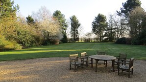 Une terrasse en gravier avec une table et des chaises en bois, entourée d’une pelouse bien entretenue et d’arbres.