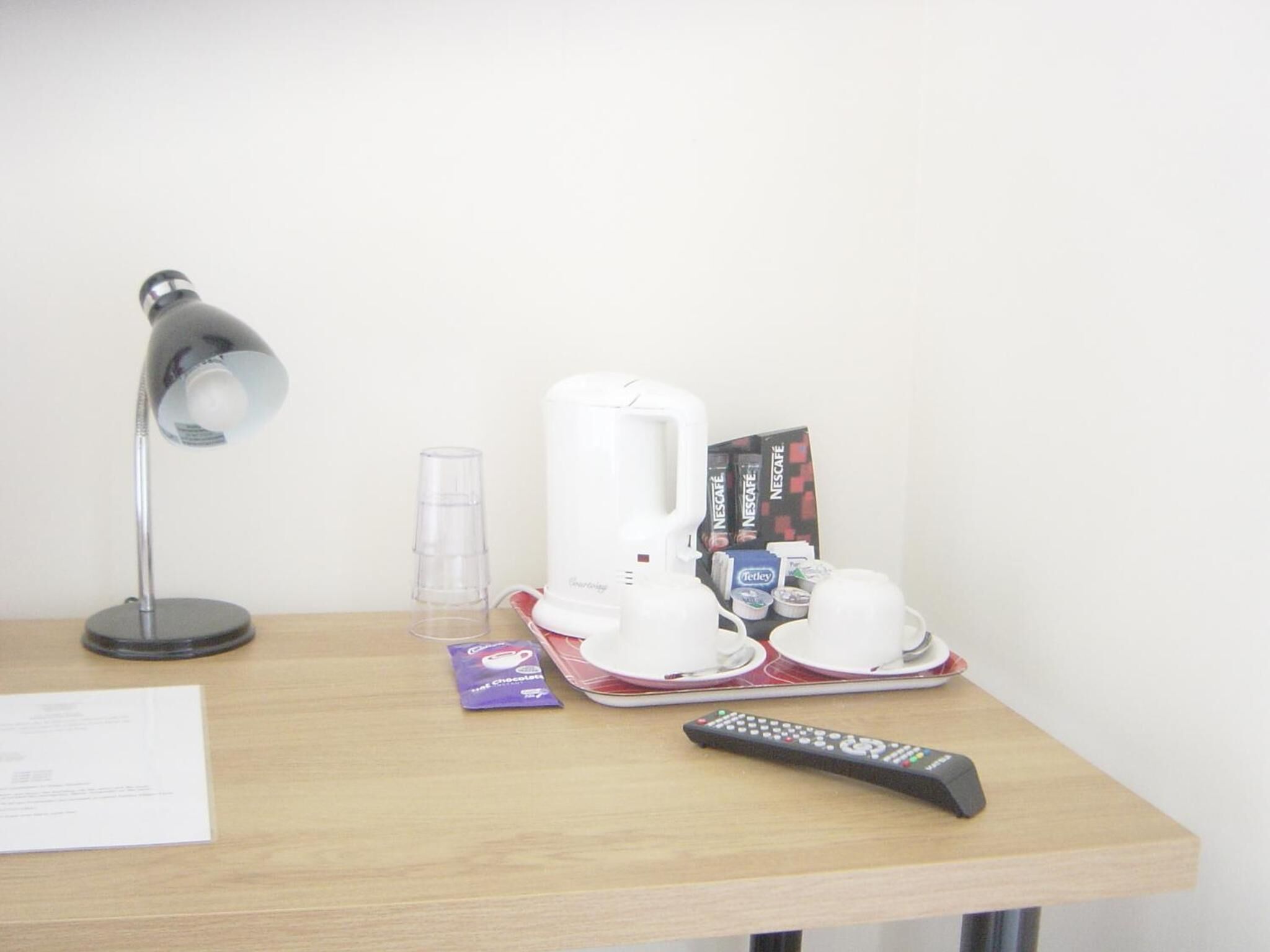 A desk with a lamp, a coffee machine, a remote control, and some coffee pods.