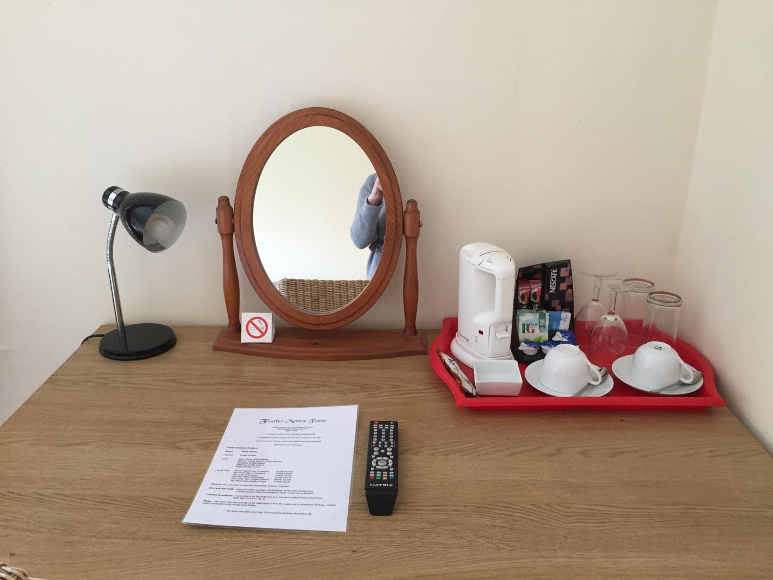 A wooden desk with a mirror, a desk lamp, a coffee maker, a tray with cups and glasses, a remote control, and a menu.