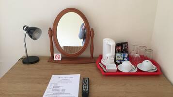 A wooden desk with a mirror, a desk lamp, a coffee maker, a tray with cups and glasses, a remote control, and a menu.