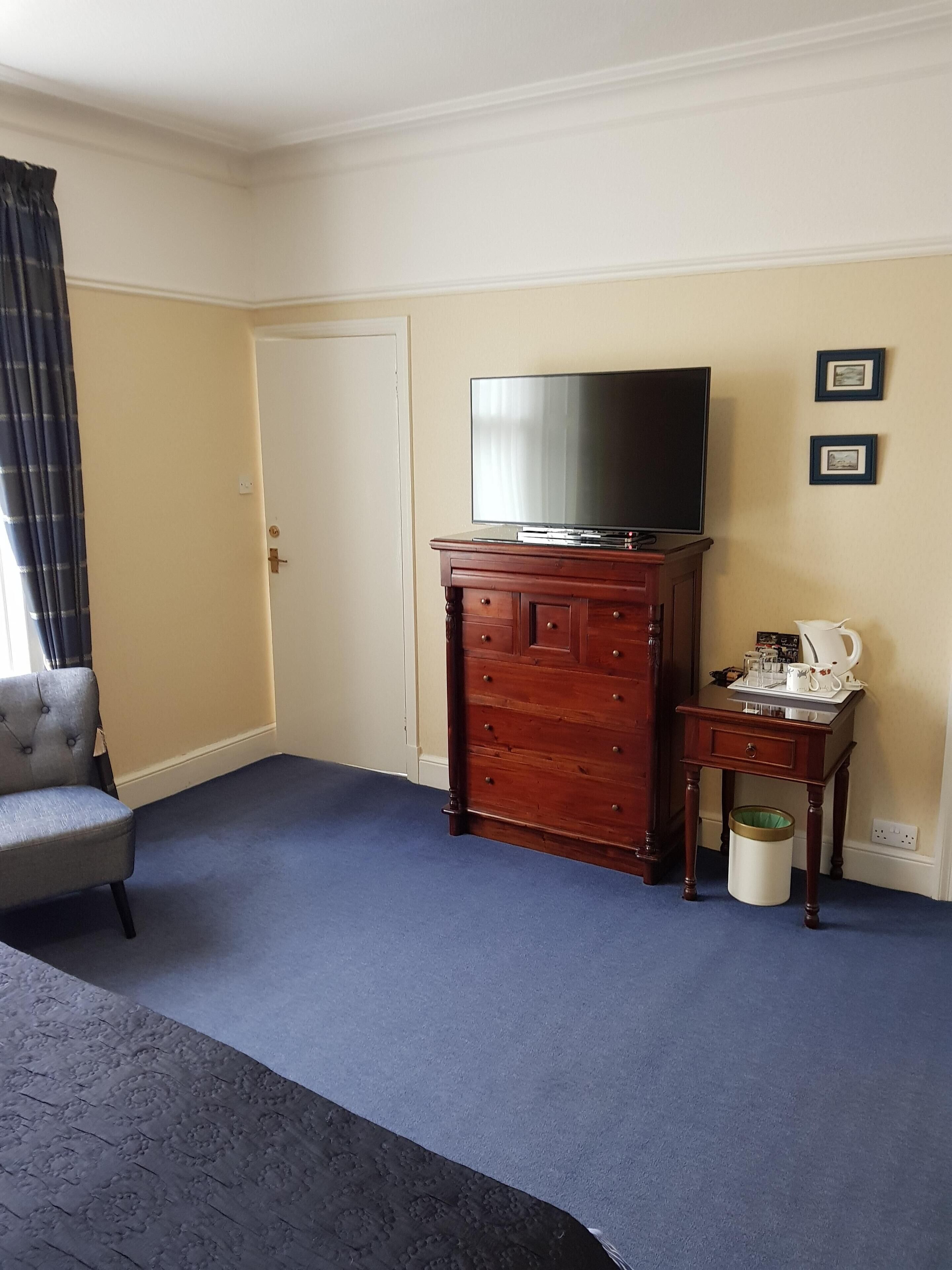 Une chambre d’hôtel avec un grand téléviseur à écran plat posé sur une commode en bois, un fauteuil gris, une petite table avec une lampe et une poubelle.
