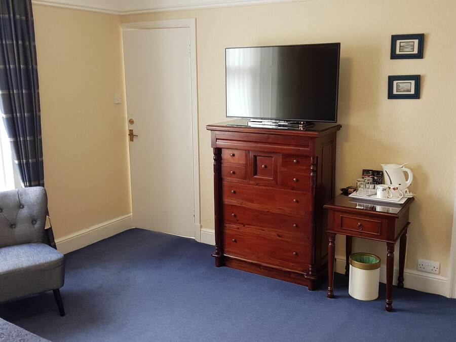 Une chambre d’hôtel avec un grand téléviseur à écran plat posé sur une commode en bois, un fauteuil gris, une petite table avec une lampe et une poubelle.
