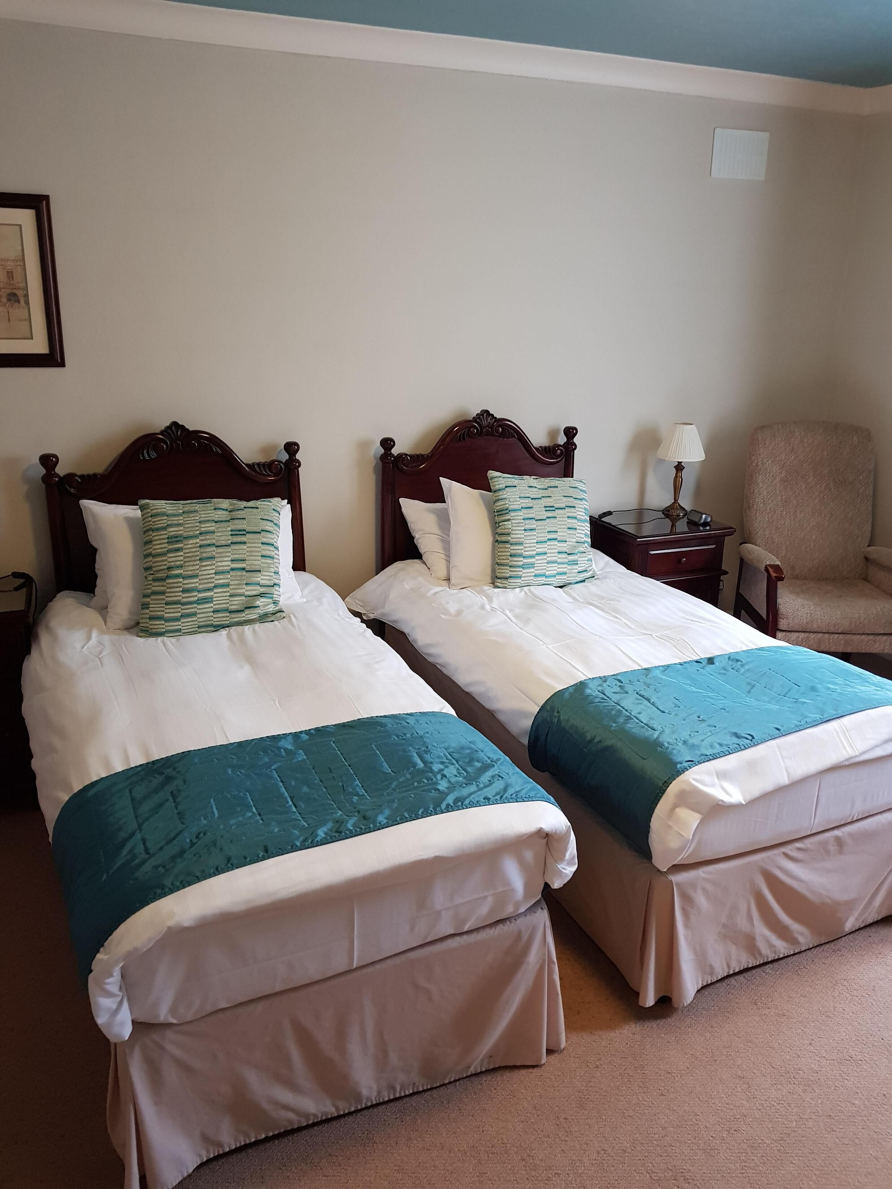 A hotel room with two beds, each with a wooden headboard, white bedding, and teal throw pillows. There is a nightstand with a lamp, a chair, and a framed picture on the wall.