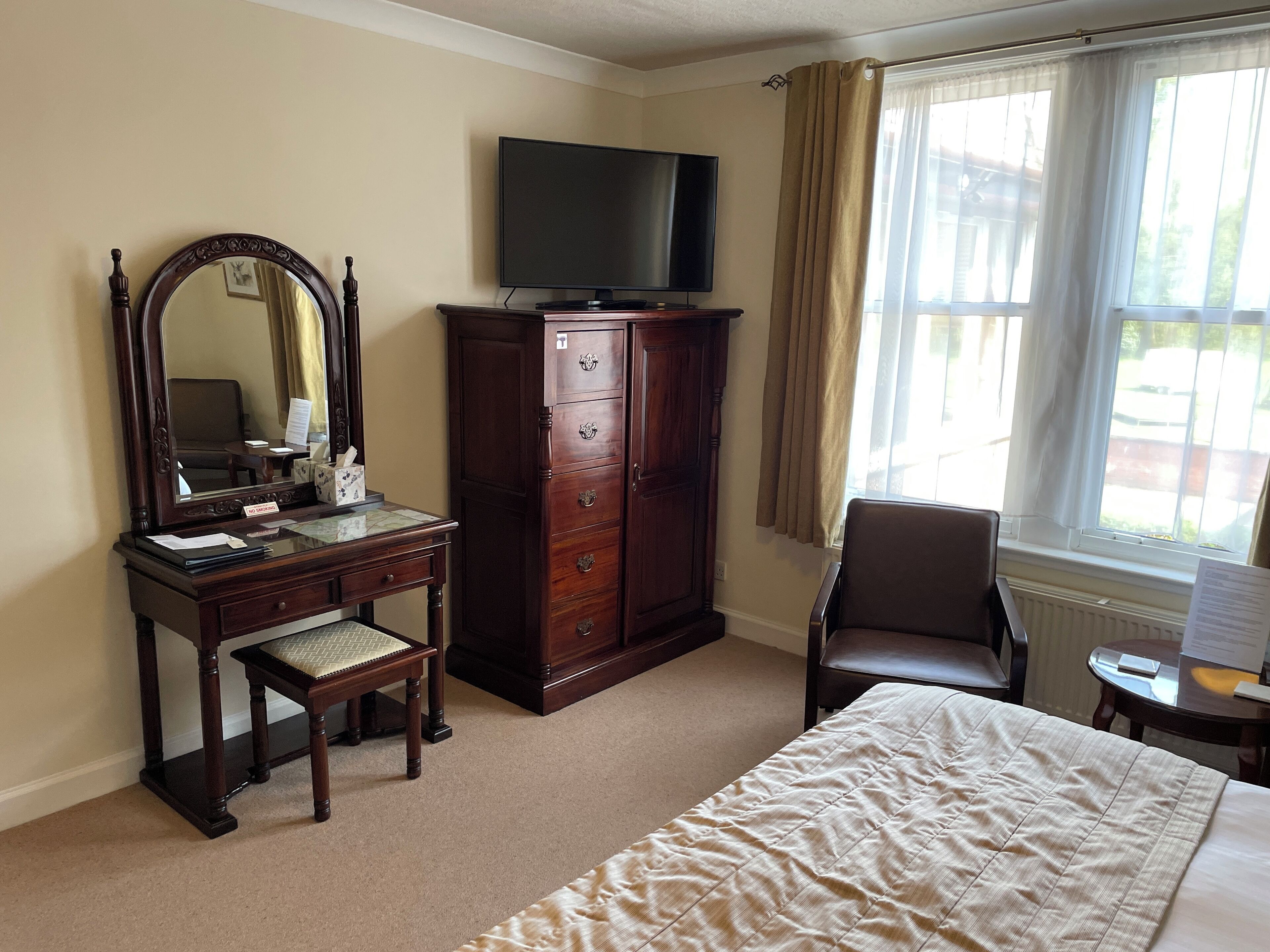 Une chambre d’hôtel avec un lit, une commode en bois, un miroir, une télévision, une chaise et une fenêtre avec des rideaux.