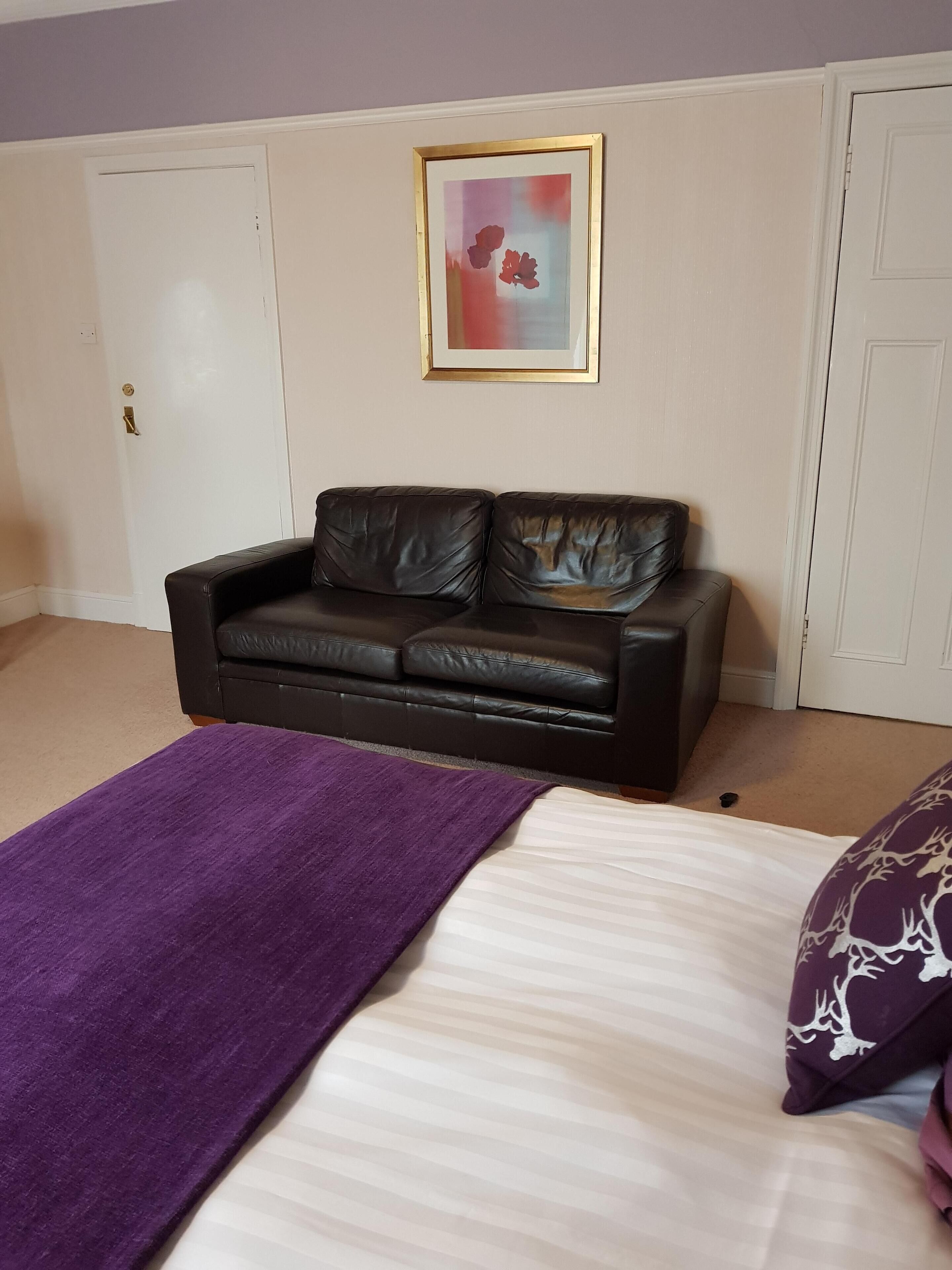 Una camera da letto con un letto, un divano in pelle nera, una coperta viola e un quadro appeso al muro.