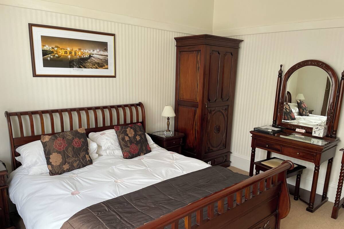 A neatly arranged bedroom with a large bed, a wooden wardrobe, a dresser with a mirror, and a framed picture on the wall.
