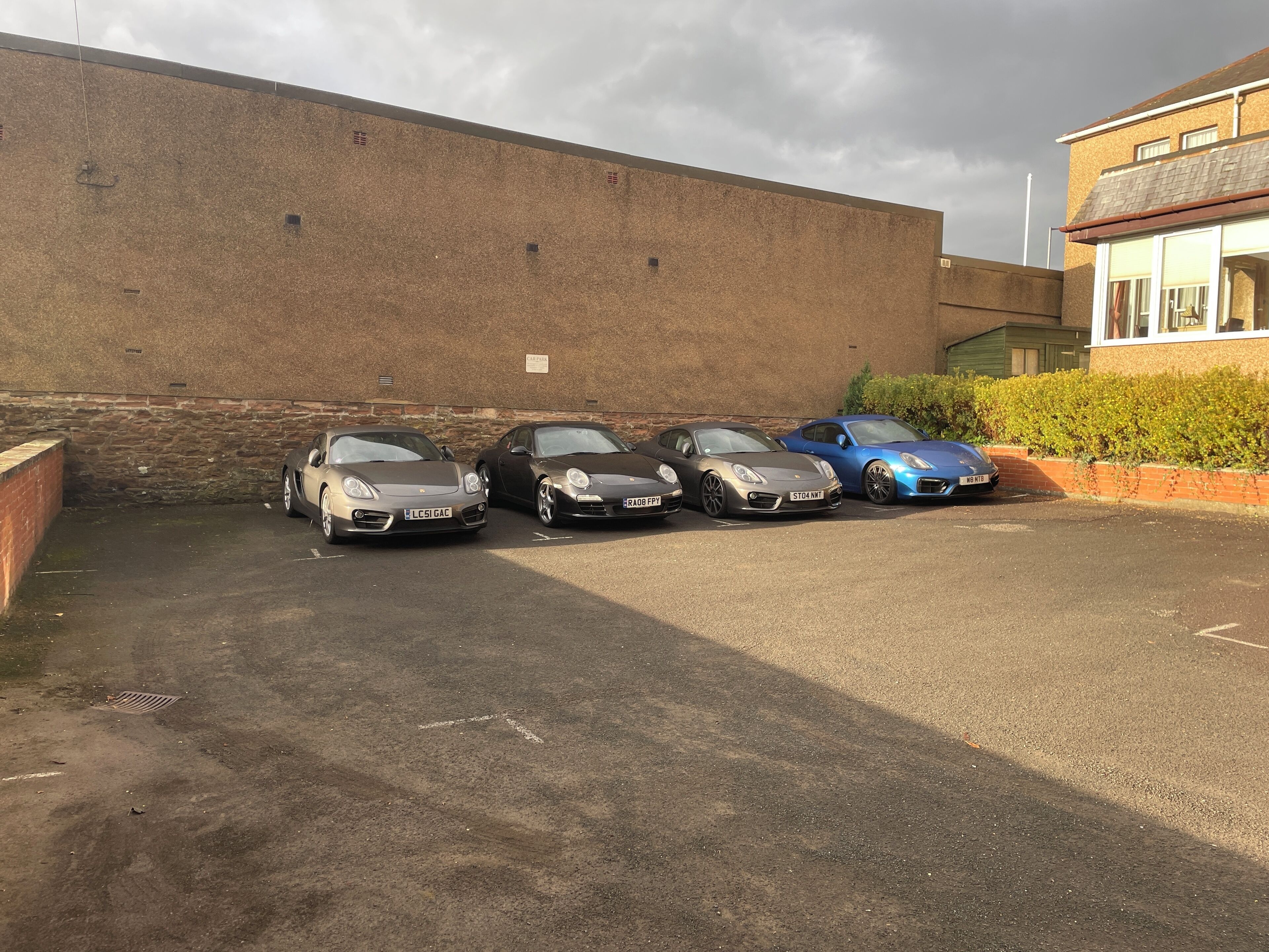 Un área de estacionamiento con cuatro coches aparcados, un muro de ladrillo y un edificio con ventanas y una puerta.