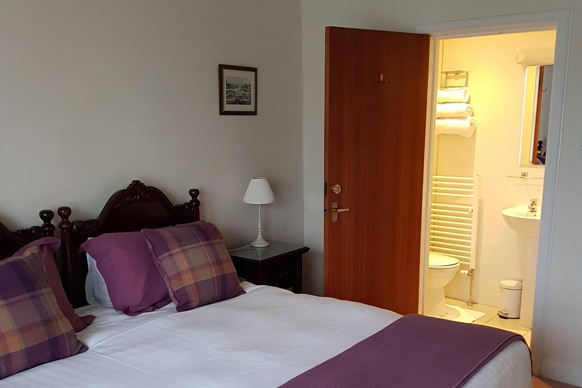 A neatly made bed with a white and purple bedspread, a wooden headboard, and two pillows. A nightstand with a white lamp is beside the bed. A wooden door leads to a bathroom with a white sink and mirror.