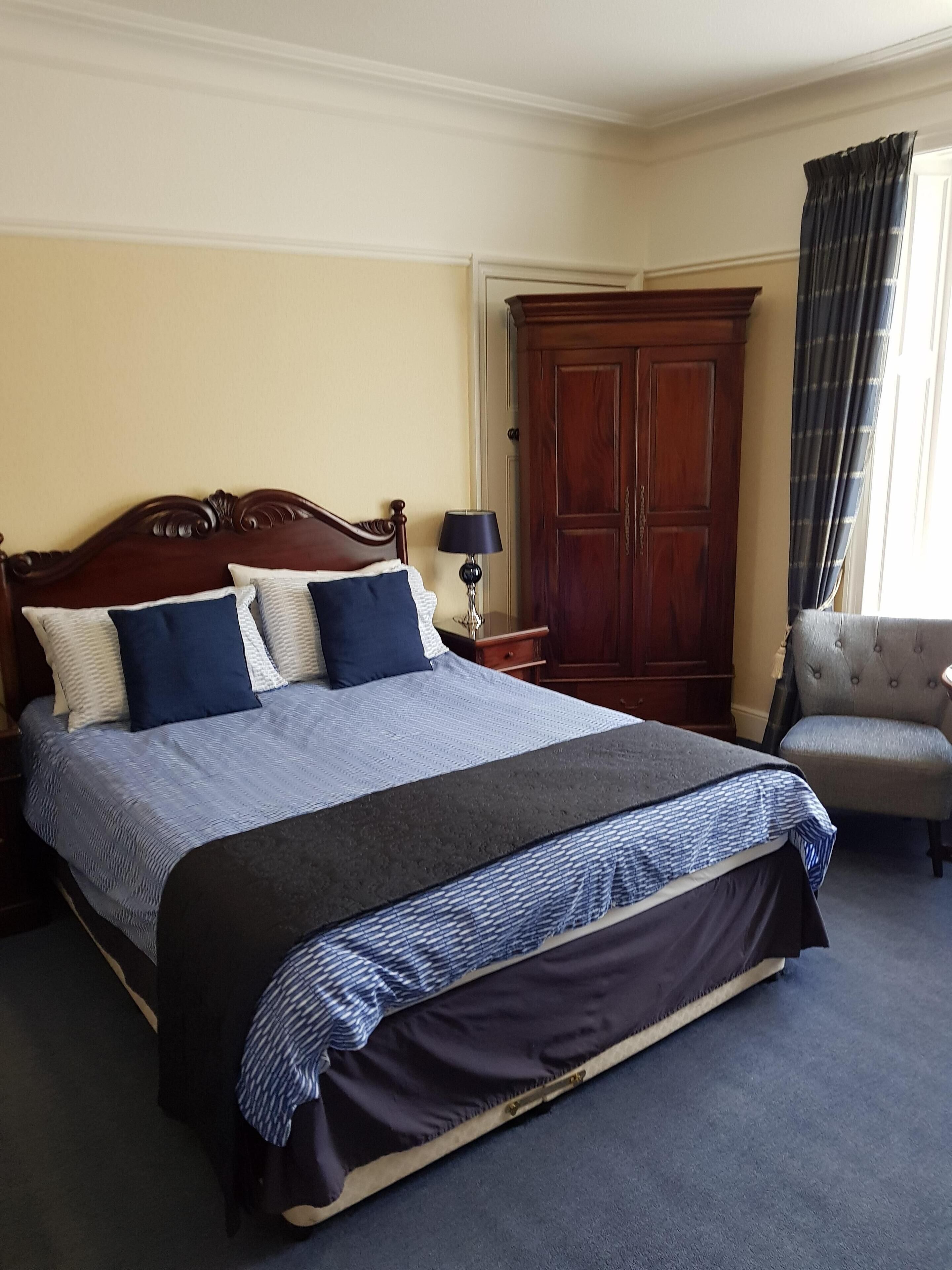 A neatly made bed with a dark headboard, a bedside table with a lamp, a wooden wardrobe, and a chair.
