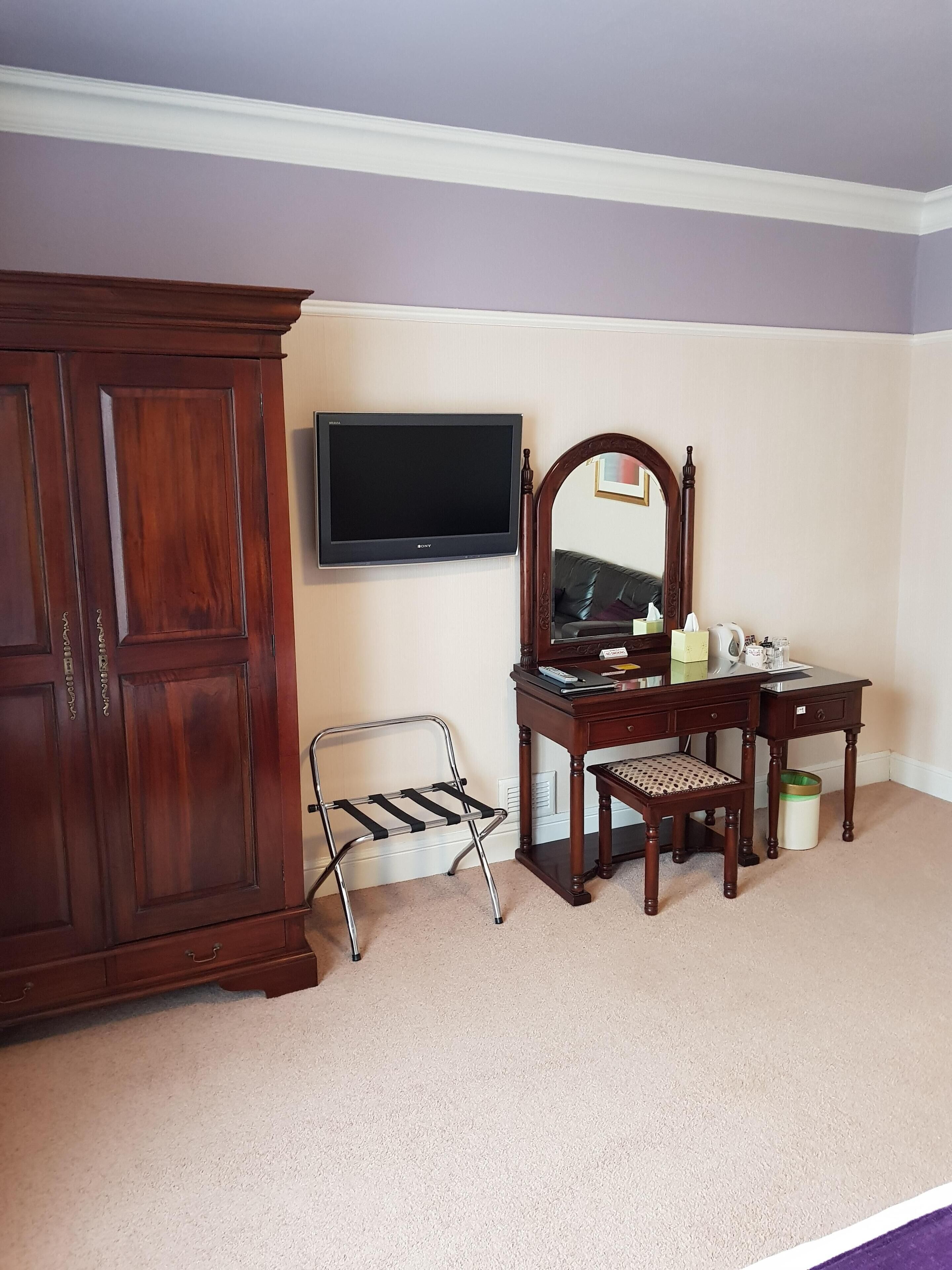 A room with a wooden wardrobe, a television mounted on the wall, a desk with a mirror, and a chair.