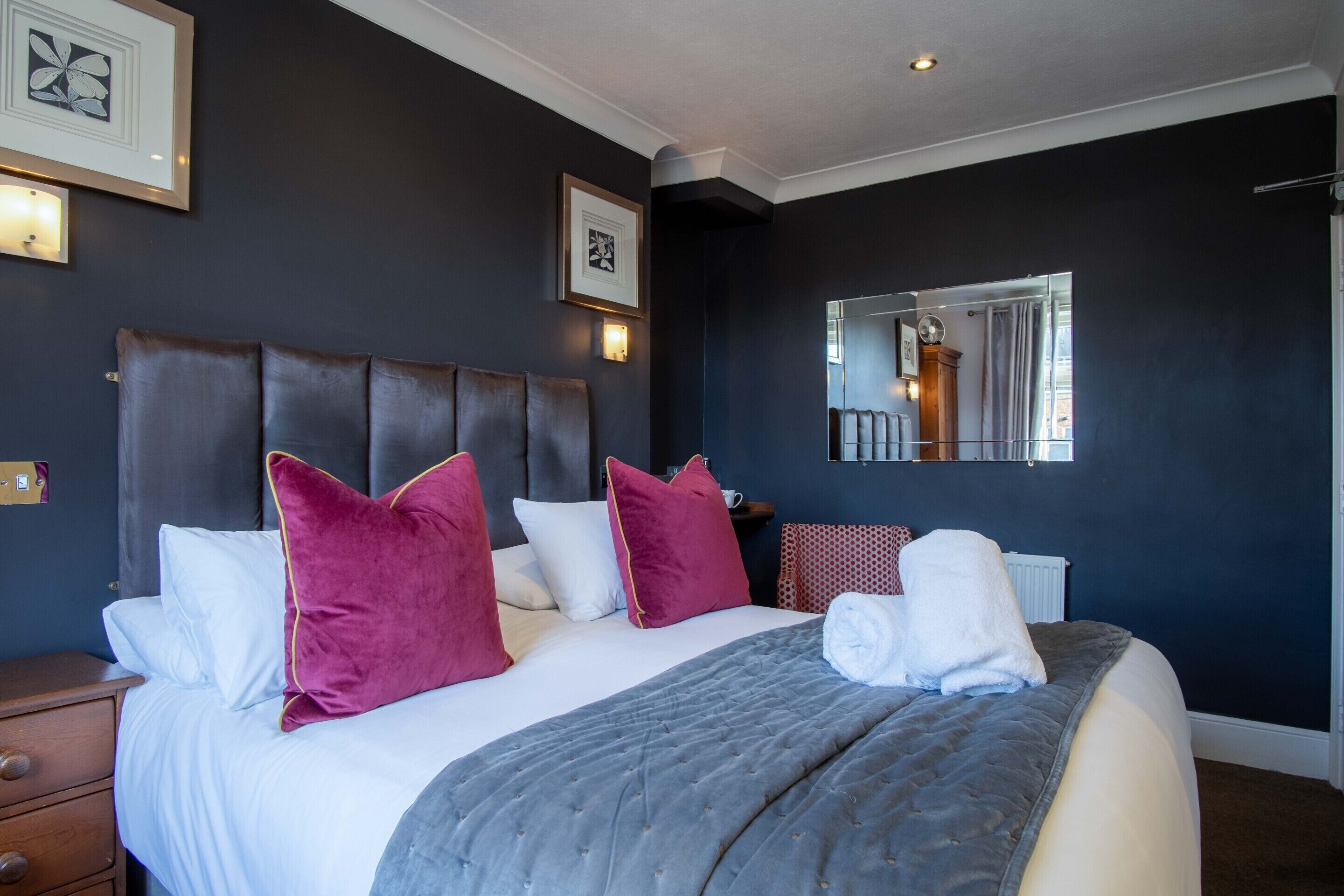 Una cama bien hecha con colcha gris, cabecera oscura y varios almohadones rojo y blanco.