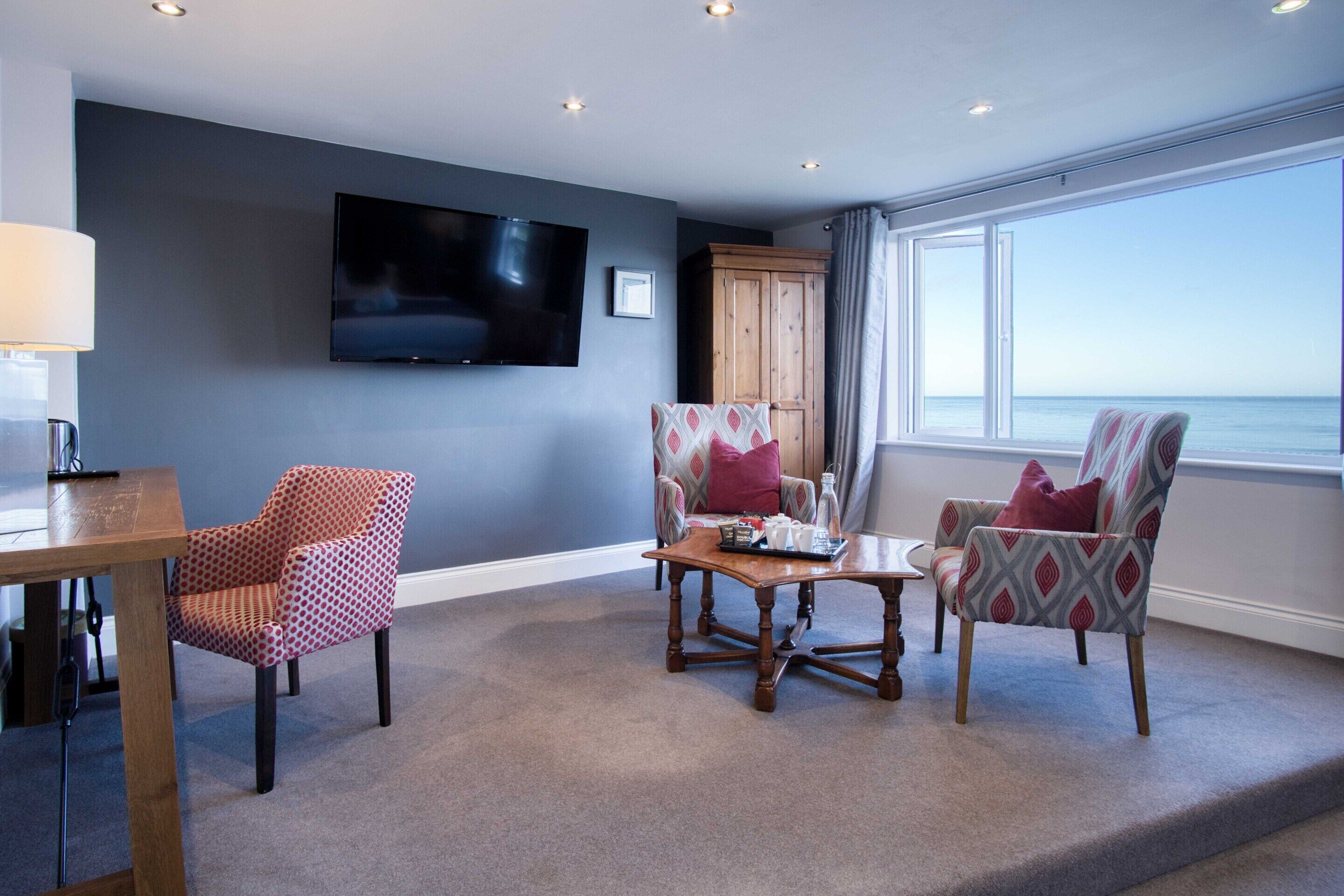 A modern living room with a flat-screen TV, two patterned armchairs, a wooden coffee table, and a view of the sea through large windows.