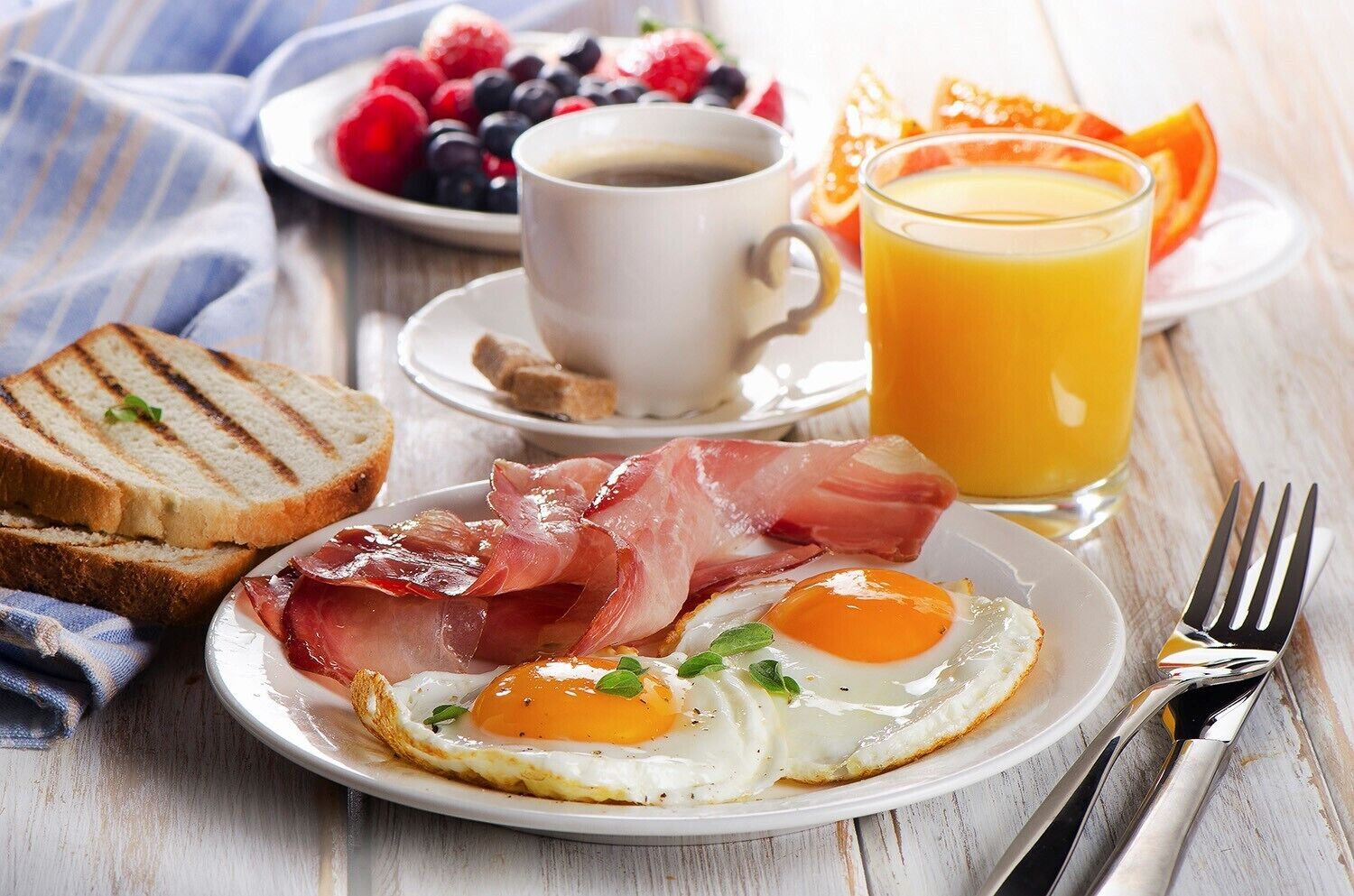 Une assiette de petit-déjeuner avec des œufs, du jambon de Parme et des toasts, une tasse de café et un verre de jus d’orange, le tout posé sur une table en bois.