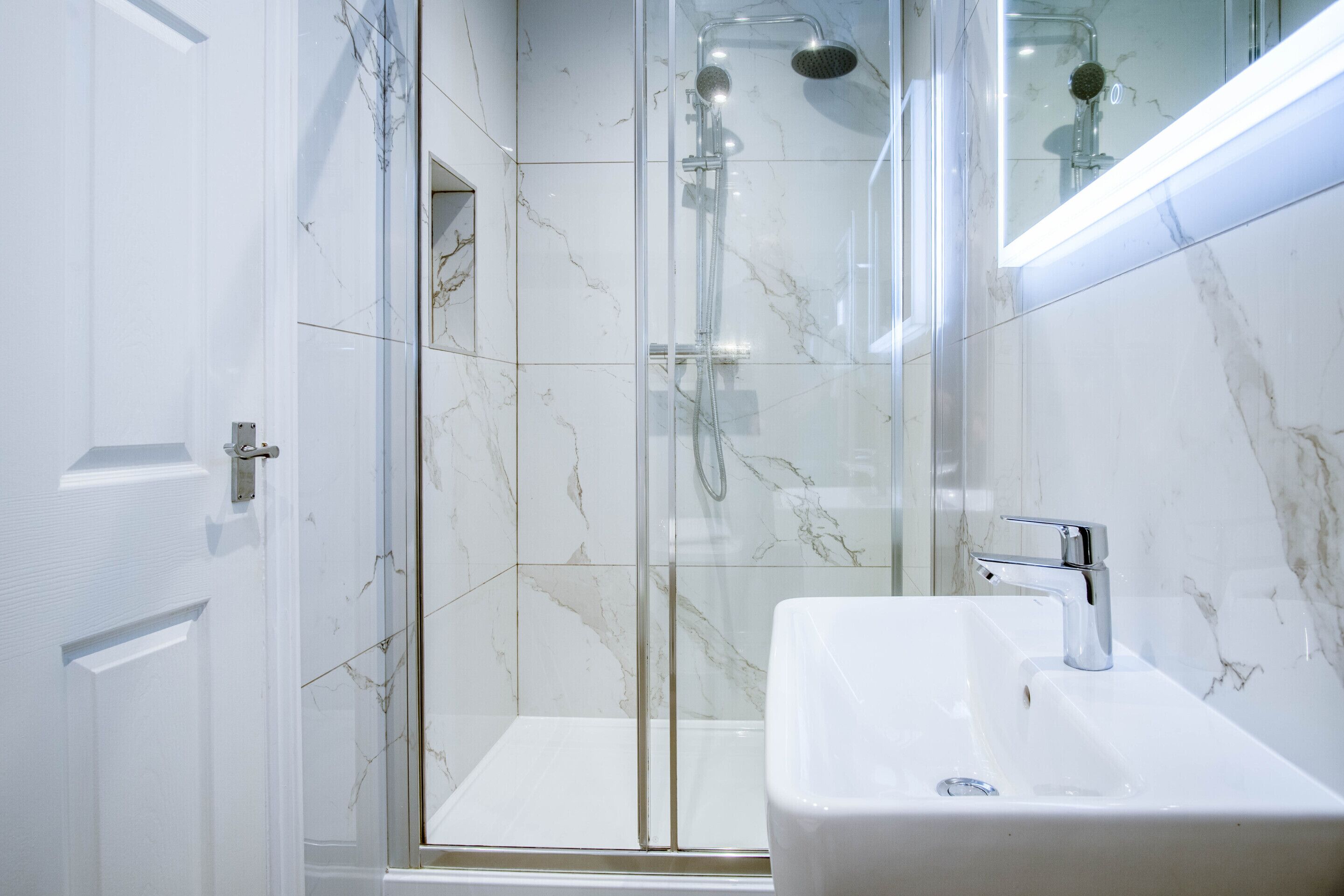 Un baño moderno con ducha de vidrio, lavamanos blanco y azulejos de mármol.
