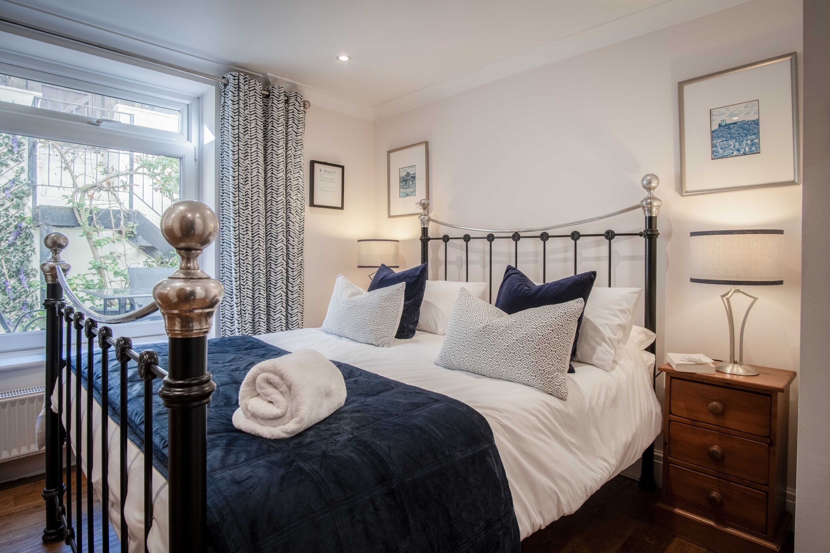 Un lit bien fait, avec une couette bleu foncé et du linge de lit blanc, une table de chevet avec une lampe et une fenêtre ornée de rideaux à motifs.