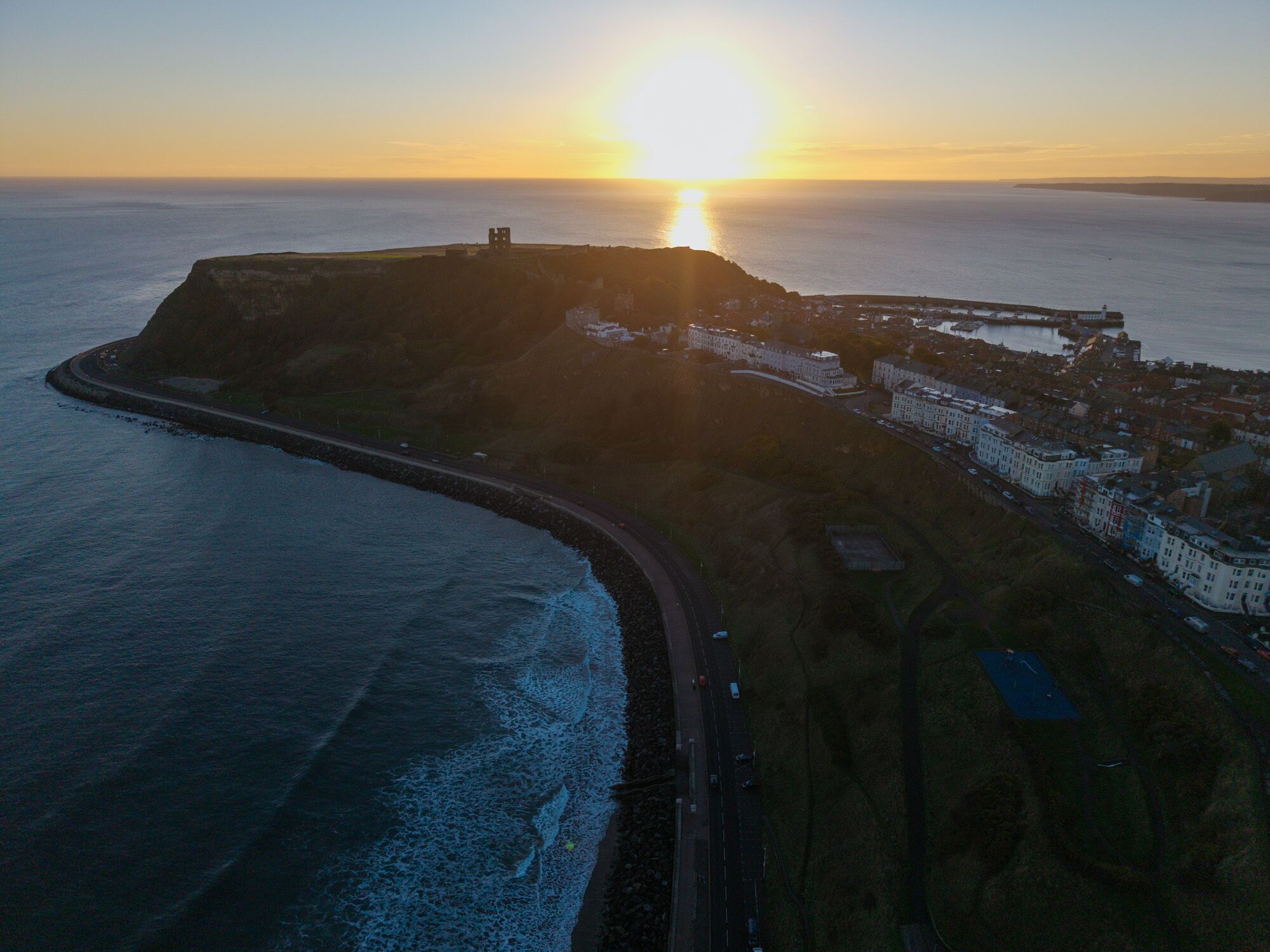 An aerial view of a coastal town with a prominent hill, a road along the coast, and buildings with a uniform architectural style.