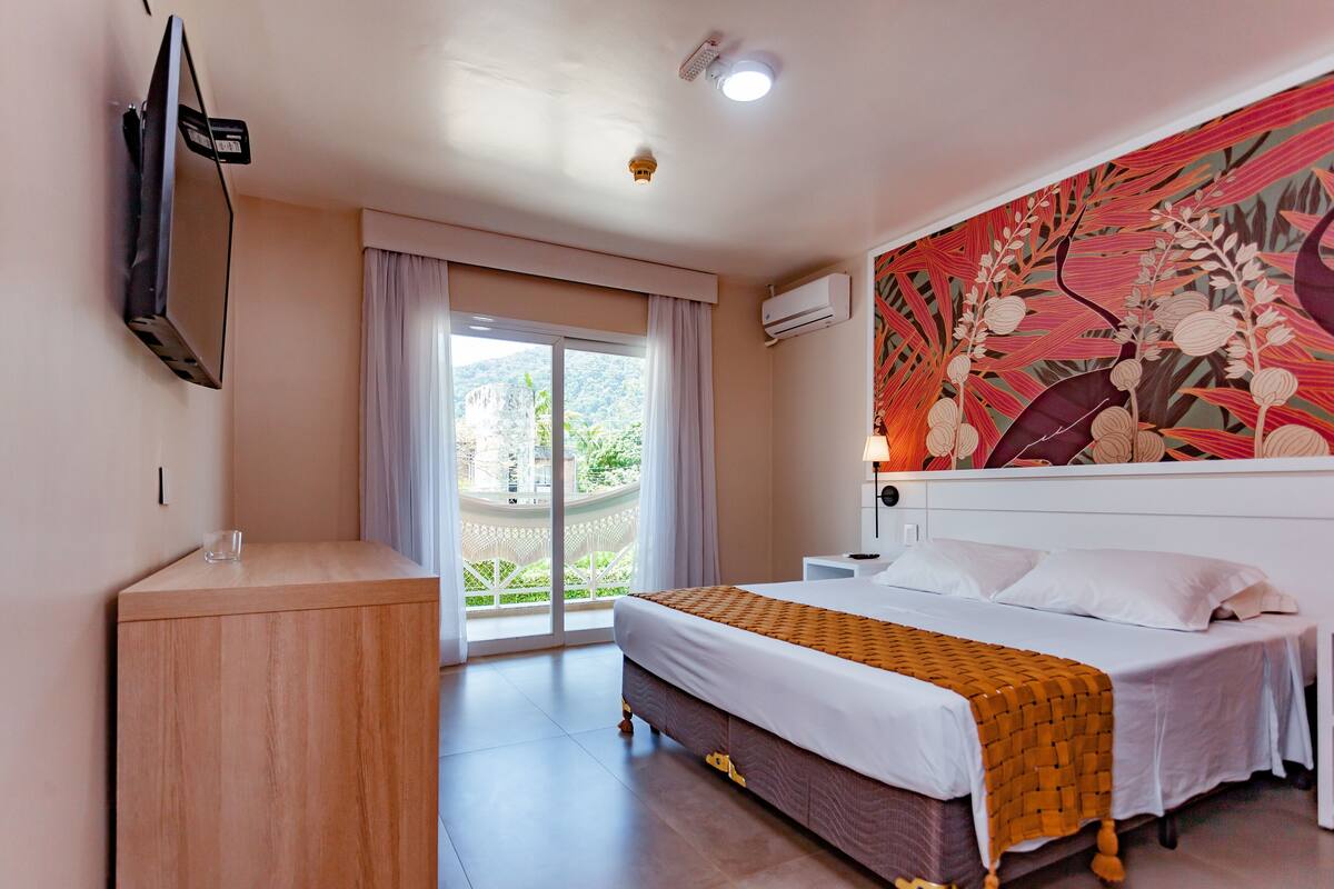 A hotel room with a bed, a TV mounted on the wall, a window with curtains, and a decorative wall mural.