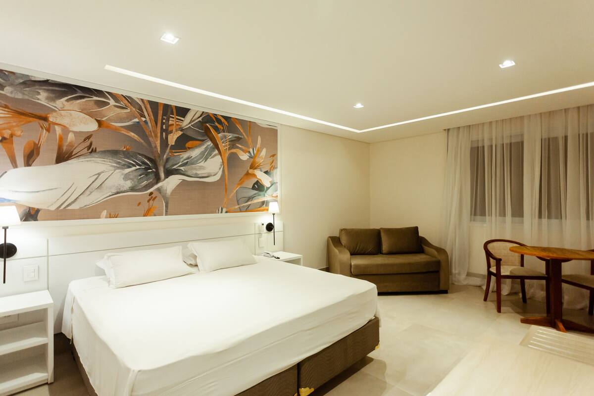 A modern hotel room with a large bed, a sofa, a dining table, and a wall-mounted painting.