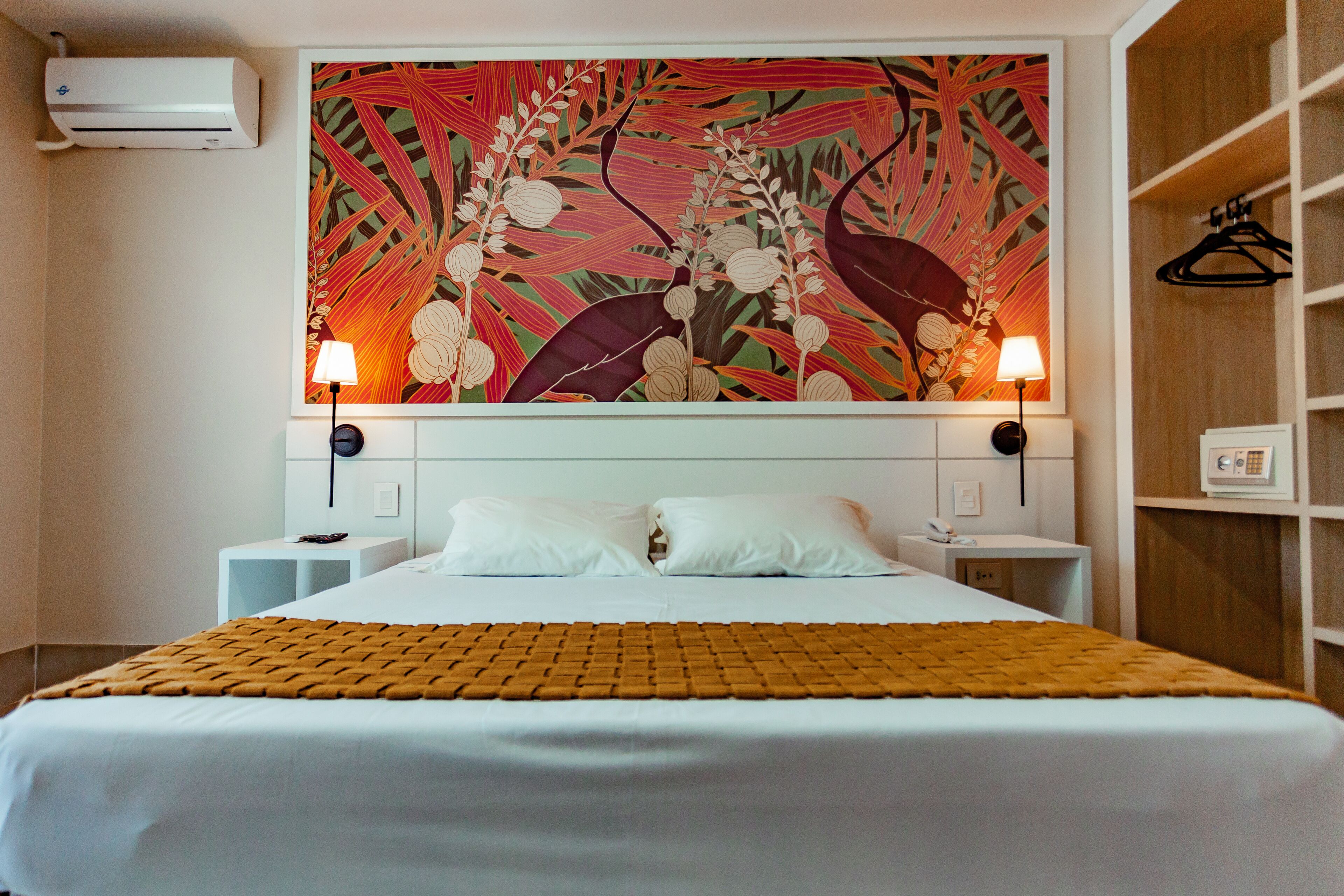 A hotel room with a large bed, two bedside tables, a wall-mounted air conditioner, and a large floral artwork above the bed.
