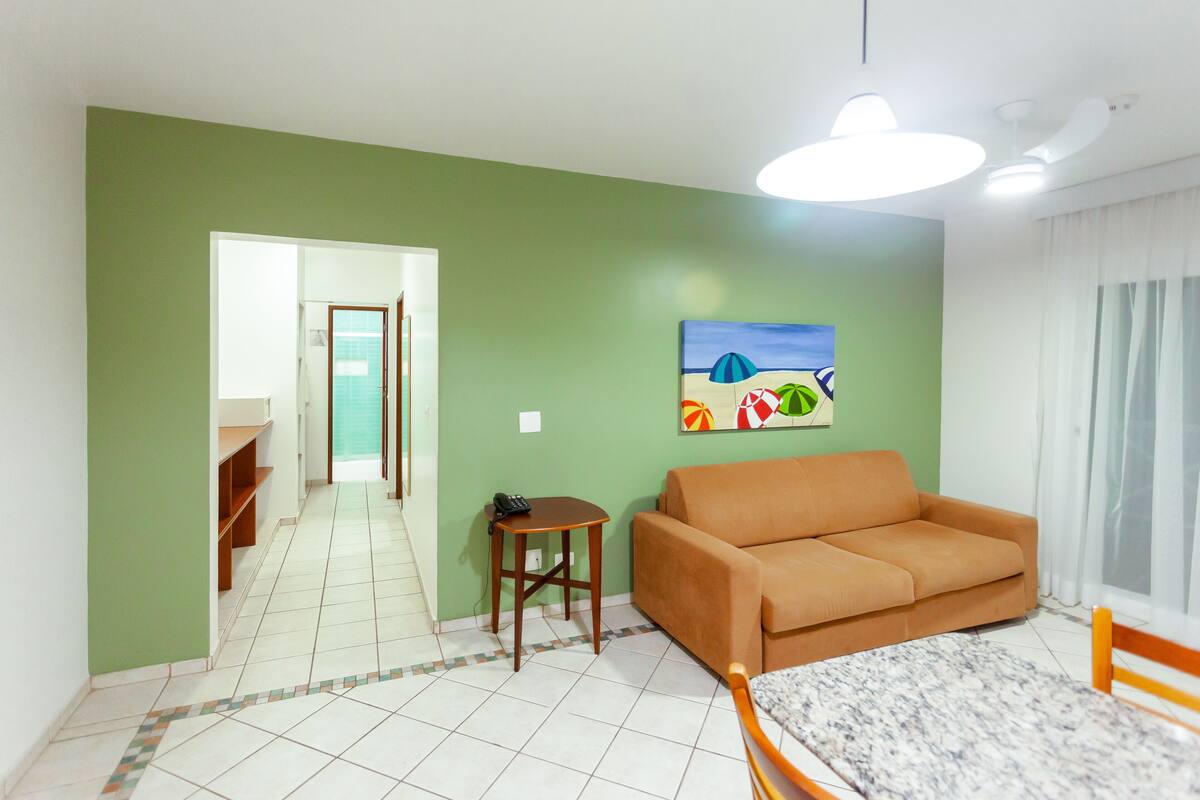 A room with a green wall, a brown sofa, a small table with a phone, a painting of a beach scene, and a tiled floor.