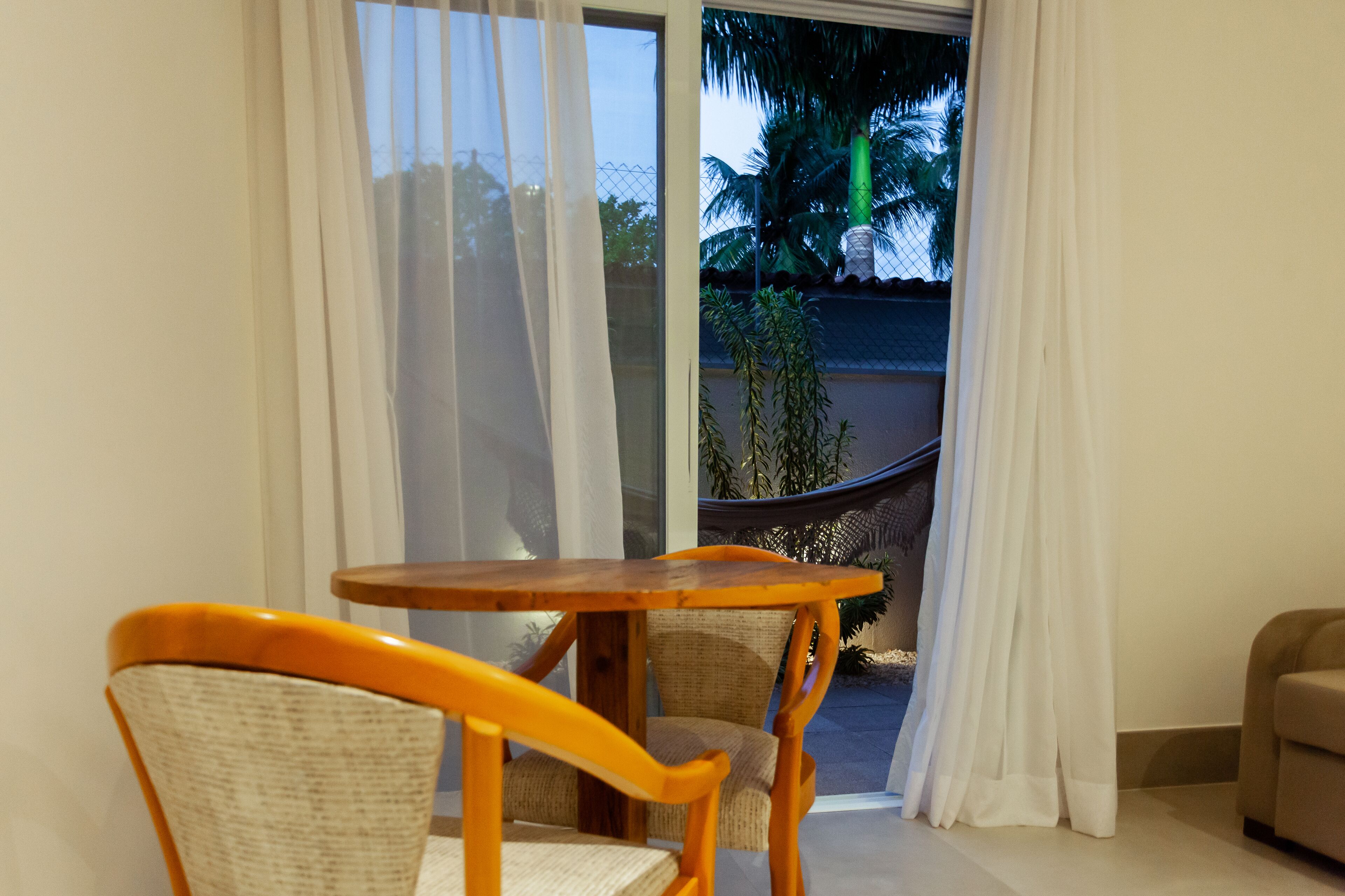 Una habitación con una mesa de madera y sillas de mimbre, una puerta corrediza de vidrio que da a un balcón con una hamaca y palmeras.