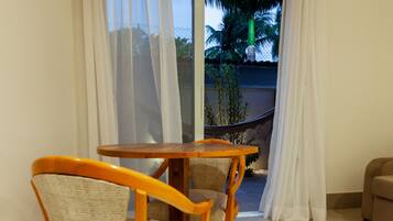Una habitación con una mesa de madera y sillas de mimbre, una puerta corrediza de vidrio que da a un balcón con una hamaca y palmeras.