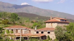 A stone building with a red-tiled roof, surrounded by greenery and mountains in the background. - Hotel Casa Rural Ropino (Candeleda)