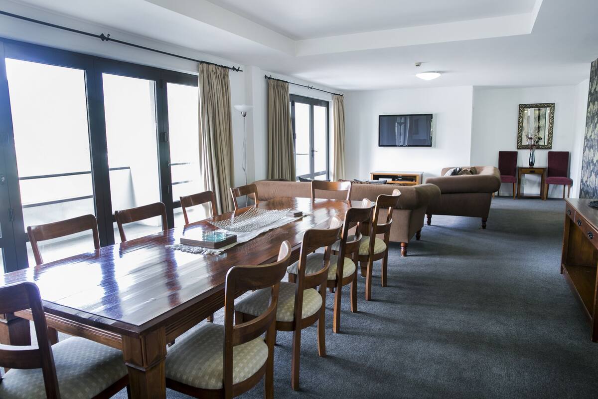 A spacious living room with a large wooden dining table, several chairs, a sofa, and a television.