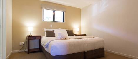 Een slaapkamer met een bed, twee nachtkastjes, een raam met jaloezieën en een vloerbedekking.
