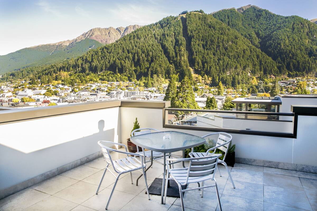 A terrace with a glass table and chairs, overlooking a town and mountains.