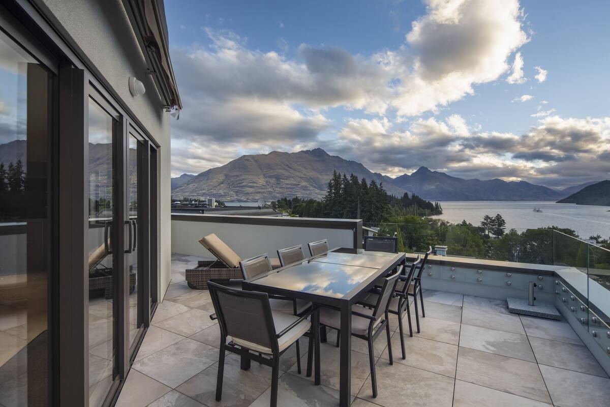 A terrace with a dining table and chairs, overlooking a mountainous landscape and a body of water.
