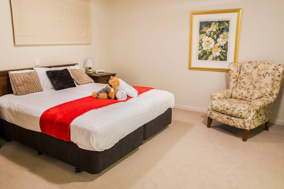 A neatly made bed with a red blanket, a floral armchair, and a framed painting on the wall.
