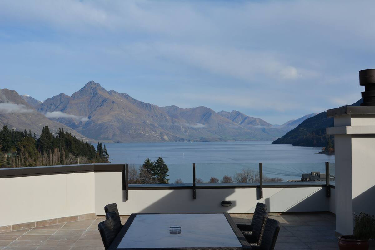A terrace with a table and chairs, overlooking a lake and mountains.