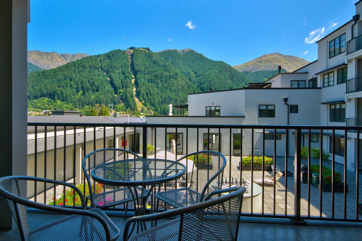 A balcony with metal chairs and tables, overlooking a mountain and a building with a white facade.
