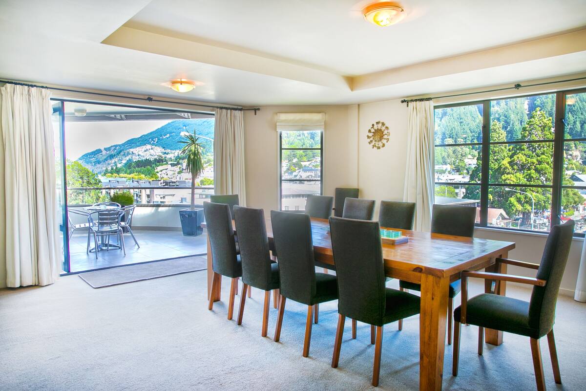 A dining area with a wooden table and chairs, large windows with a mountain view, and a balcony with a view of the mountain.