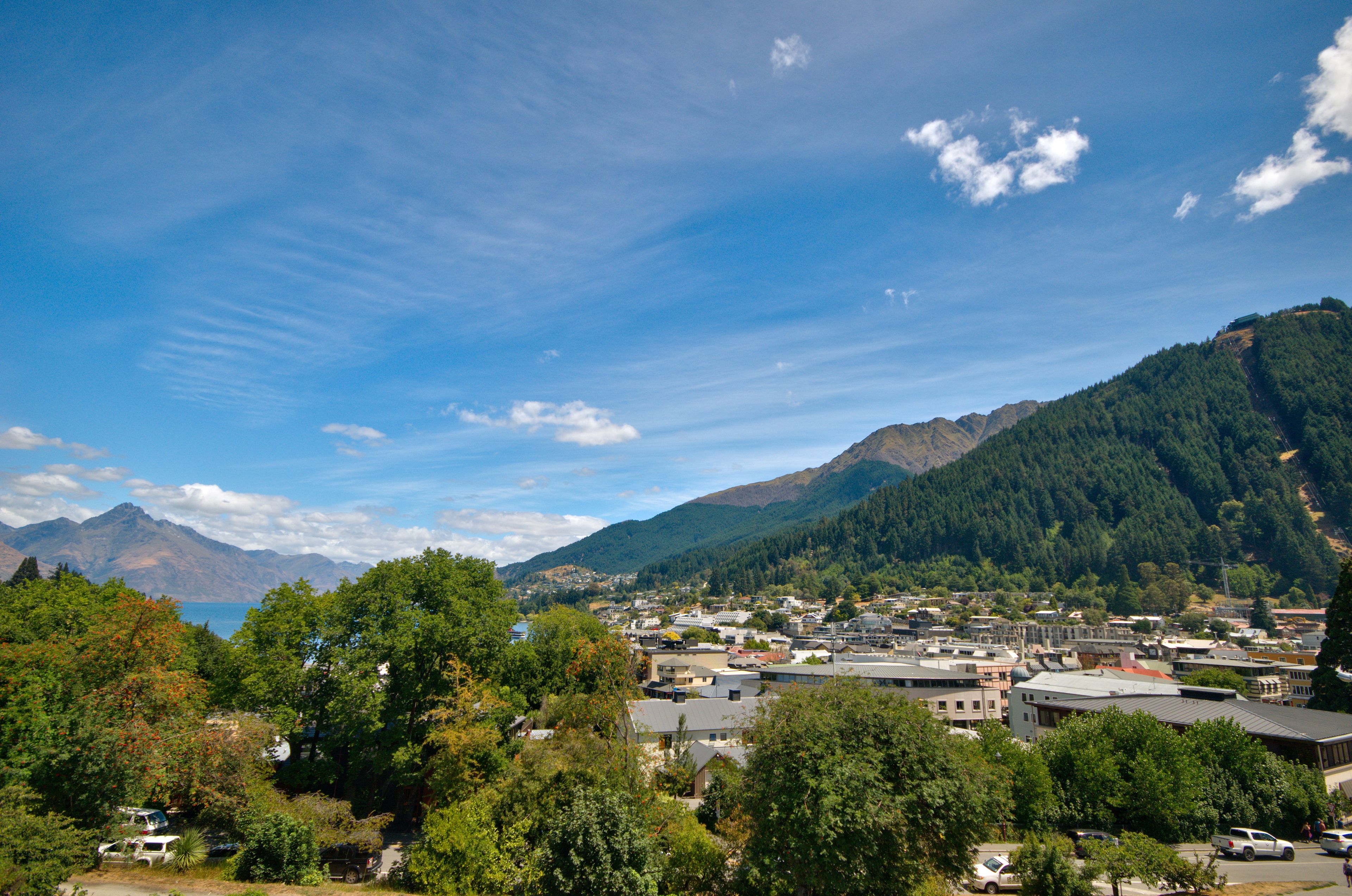 Une ville dont les bâtiments sont entourés de montagnes et d’arbres, avec un ciel dégagé et une étendue d’eau visible au loin.