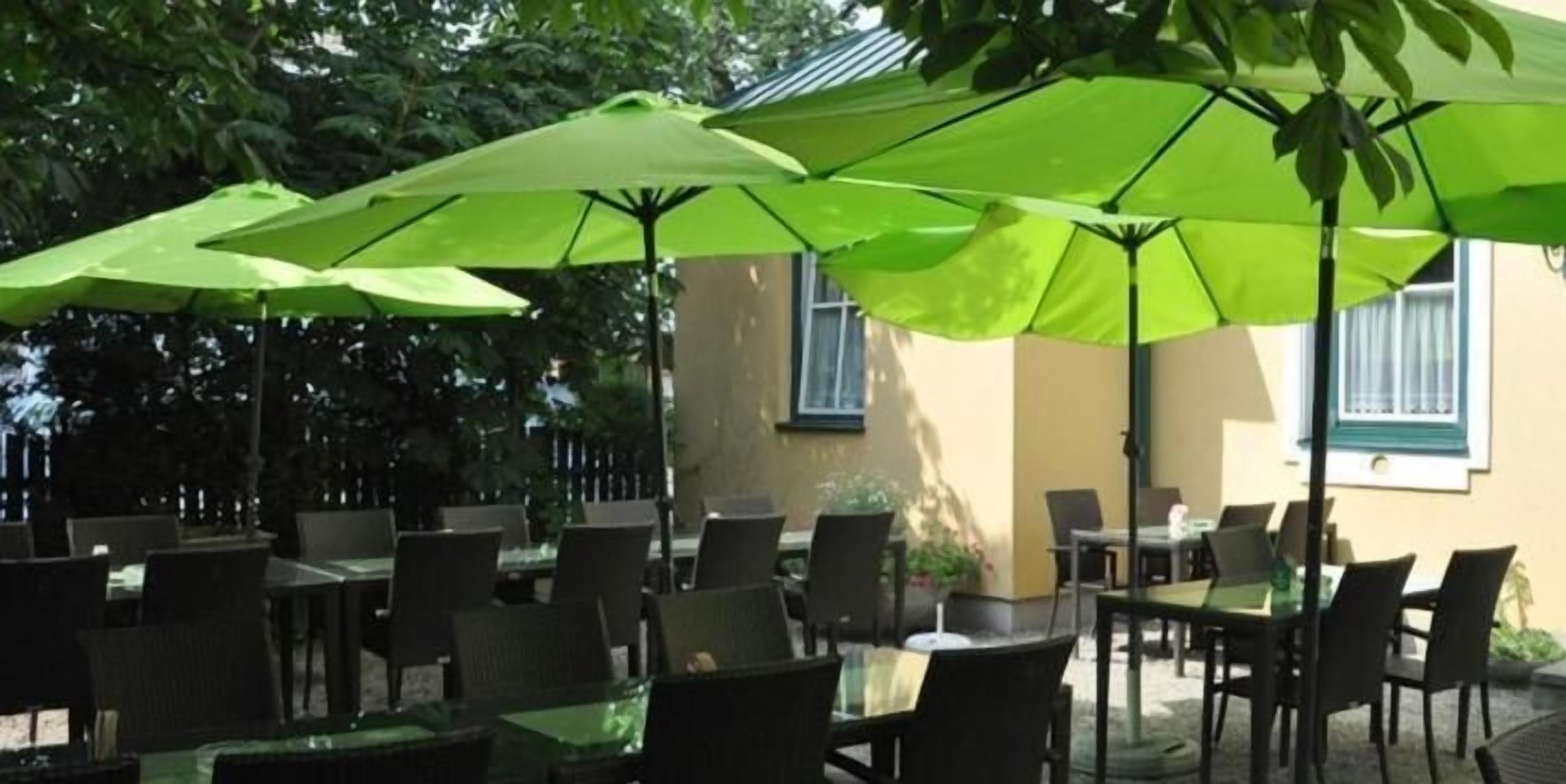 An outdoor cafe area with multiple tables and chairs, shaded by large green umbrellas.