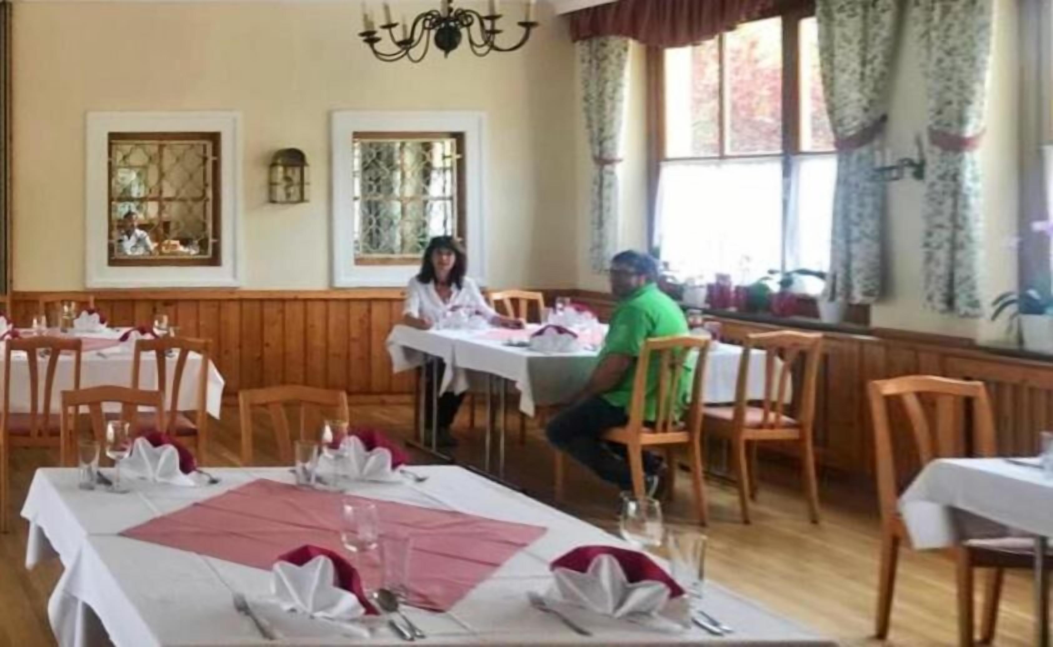 A dining area with wooden tables set for a meal, large windows with curtains, and a chandelier.