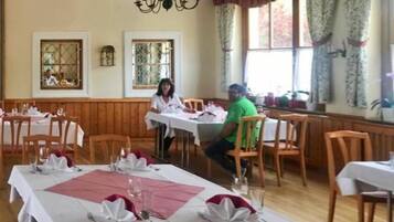 A dining area with wooden tables set for a meal, large windows with curtains, and a chandelier.