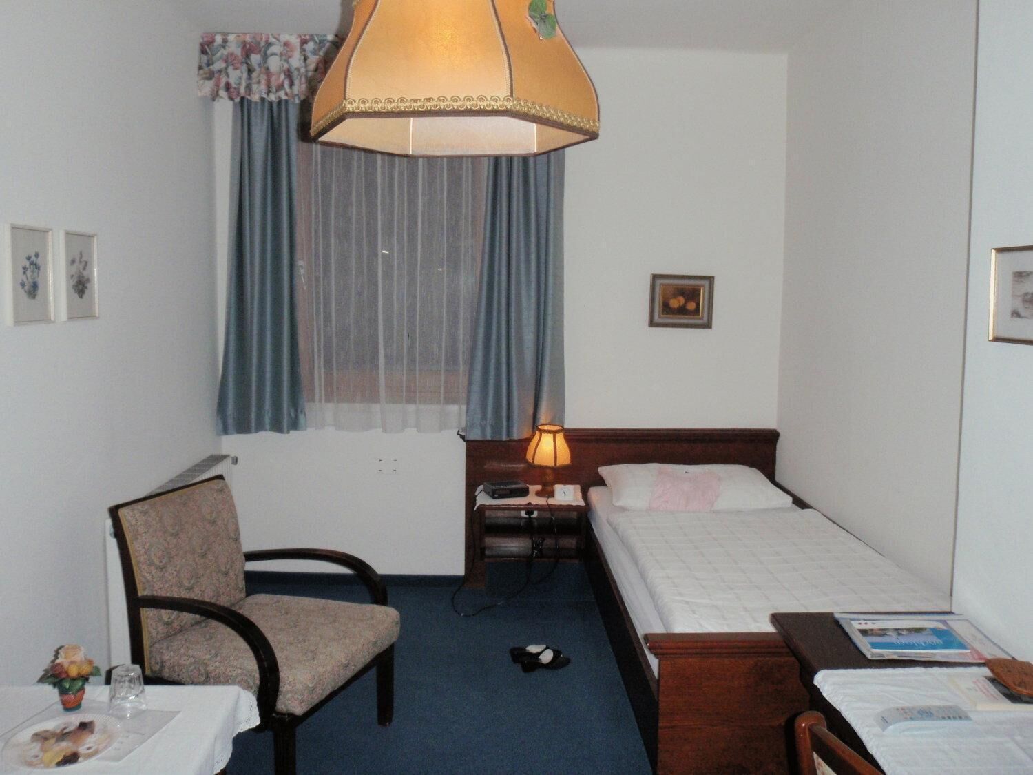 A hotel room with a single bed, a chair, a small table with a vase of flowers, a lamp, and a window with curtains.