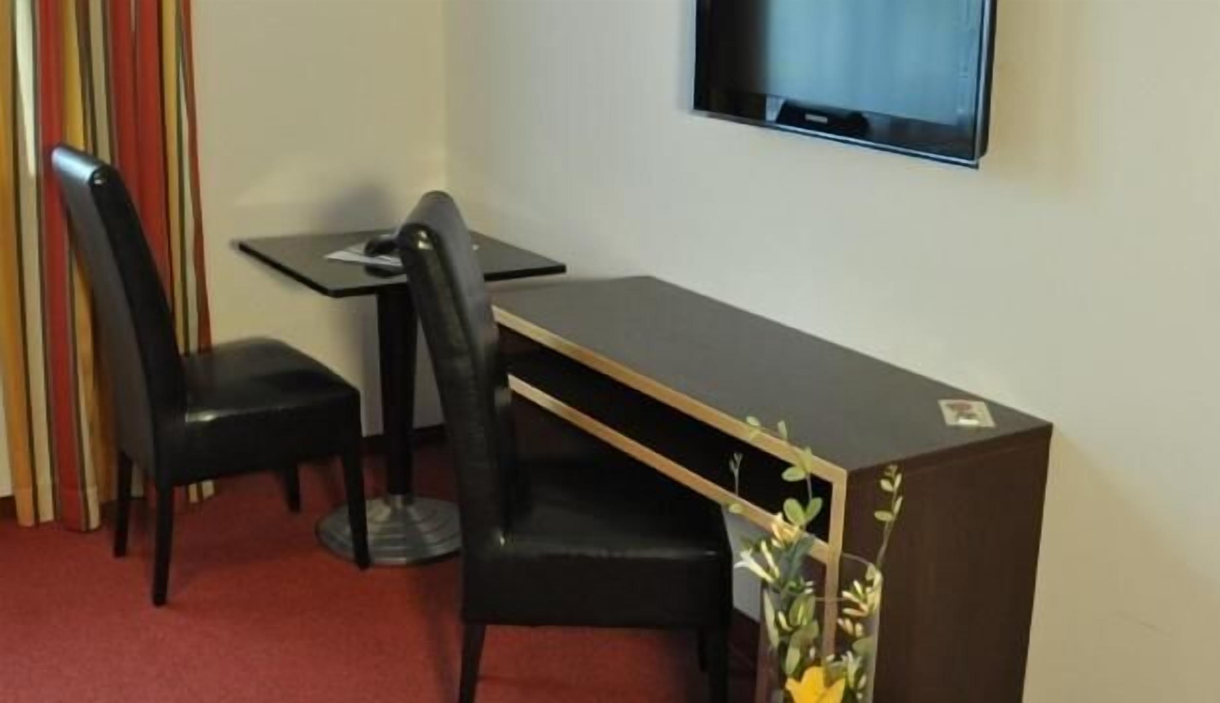 A small room with a desk, two chairs, a television mounted on the wall, and a striped curtain.