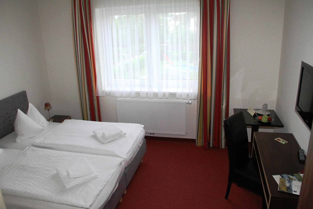 Habitación de hotel con dos camas, un escritorio, una silla, televisión y ventana con cortinas.
