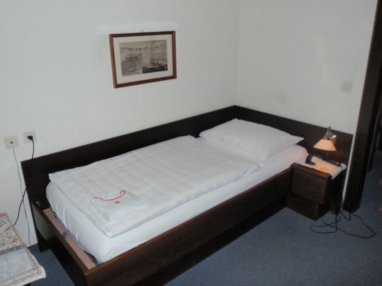 A single bed with a wooden frame, white bedding, and a bedside table with a lamp.
